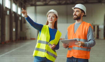 Successful workers, engineers, wearing protective workwear and safety helmets, using digital tablet, attractive woman pointing with hand, standing in warehouse. Logistics, teamwork concept